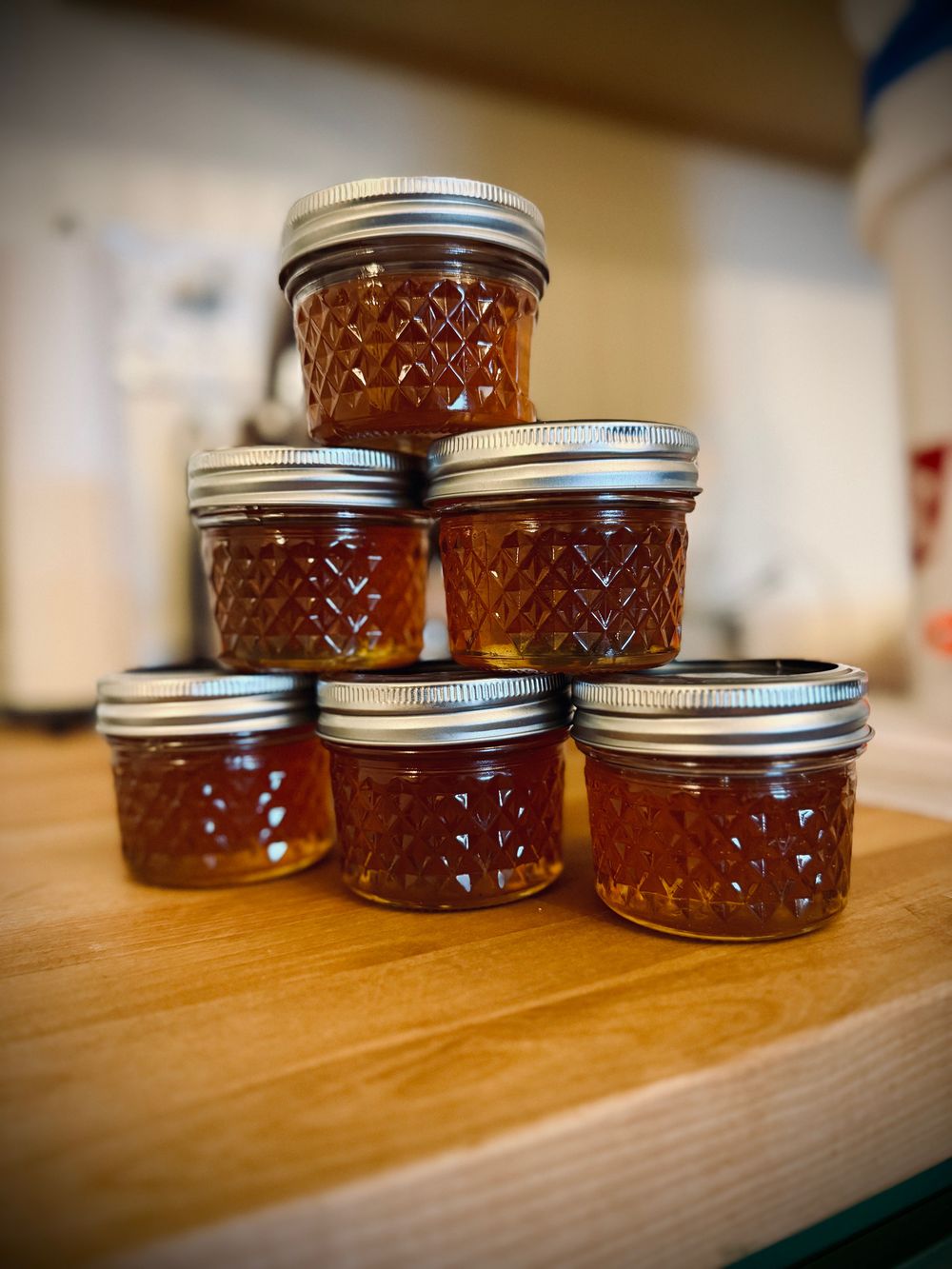 Six small quilted glass jars of amber honey stacked in a pyramid on a wooden counter